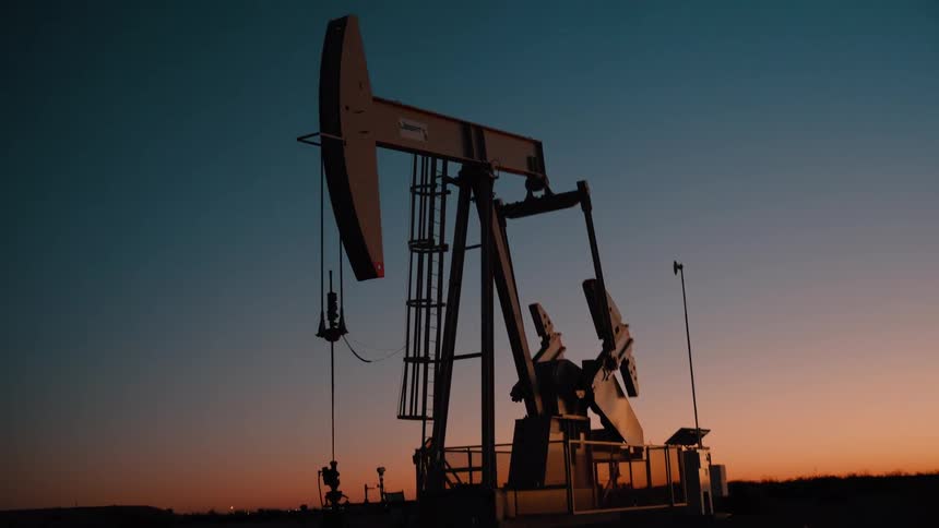 A metal pump jack silhouette moves up and down against a dark orange and blue sky during a quiet sunset in an industrial area