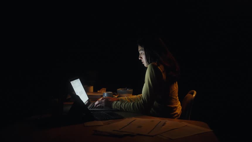 A woman wearing a yellow long sleeve shirt sits at a desk in a dark room and types on a laptop while a tablet and papers rest nearby under a soft light source