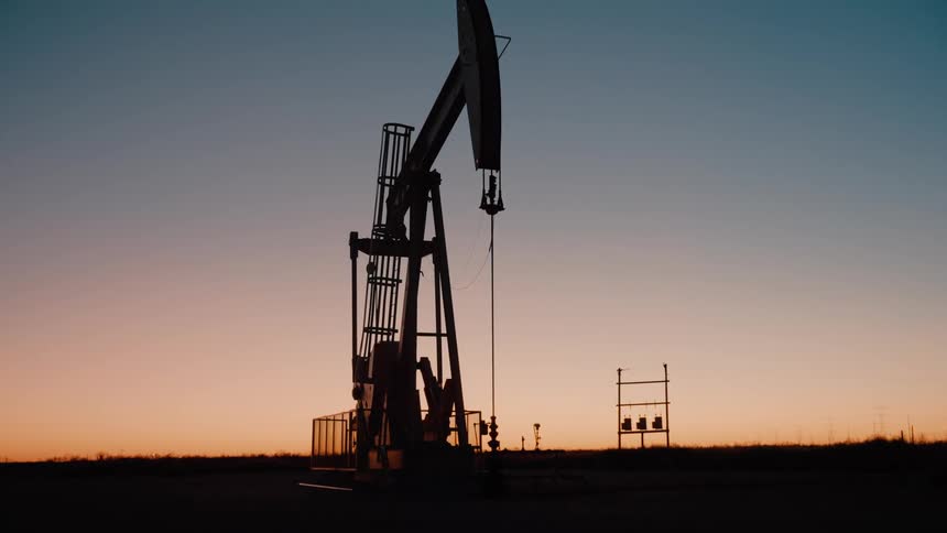 A solitary oil pumpjack operates rhythmically against a darkening sunset its mechanical silhouette standing tall as the golden horizon fades into a deep blue evening sky across the vast open plains
