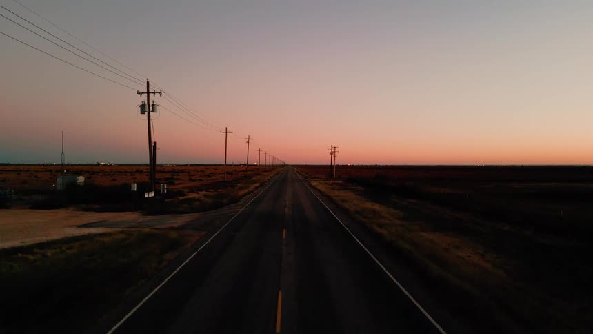 A long straight asphalt road stretches toward a distant horizon under a clear orange and purple twilight sky with wooden utility poles lining the side of the road and flat dark fields on either side