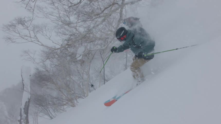 Two skiers in dark gear descend a steep mountain slope carving through deep powdery snow that kicks up thick white clouds as they navigate between bare snowladen trees during a heavy winter storm