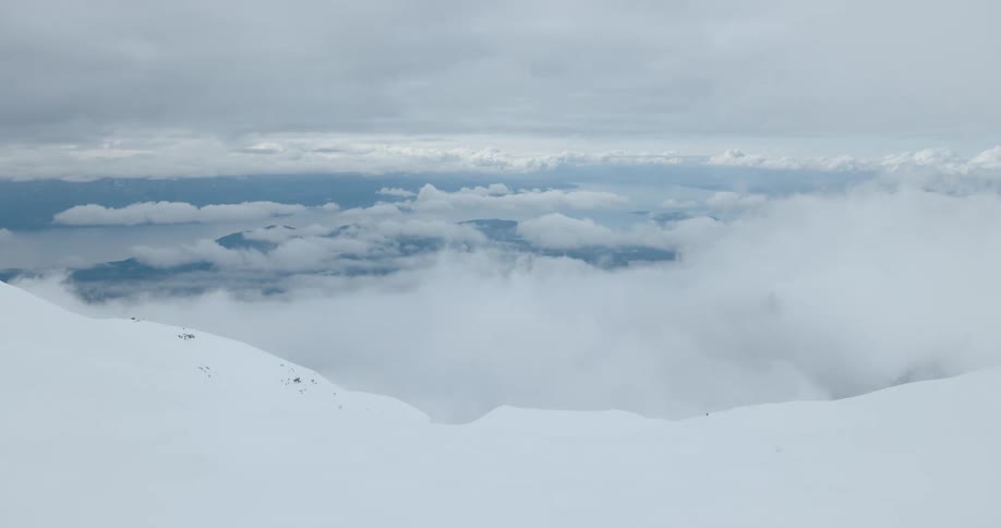 A wide shot from a snowy mountain peak shows clouds filling the valley below with distant islands and water visible on the horizon