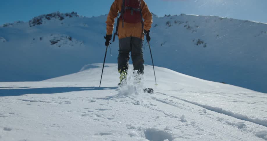 A person wearing an orange jacket and yellow boots walks away while skiing up a snowy mountain slope under a clear blue sky