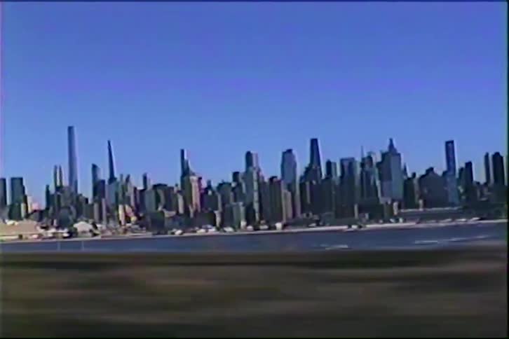 A distant view of a city skyline with many tall skyscrapers stands across a body of water under a clear blue sky as bare trees pass by in the foreground