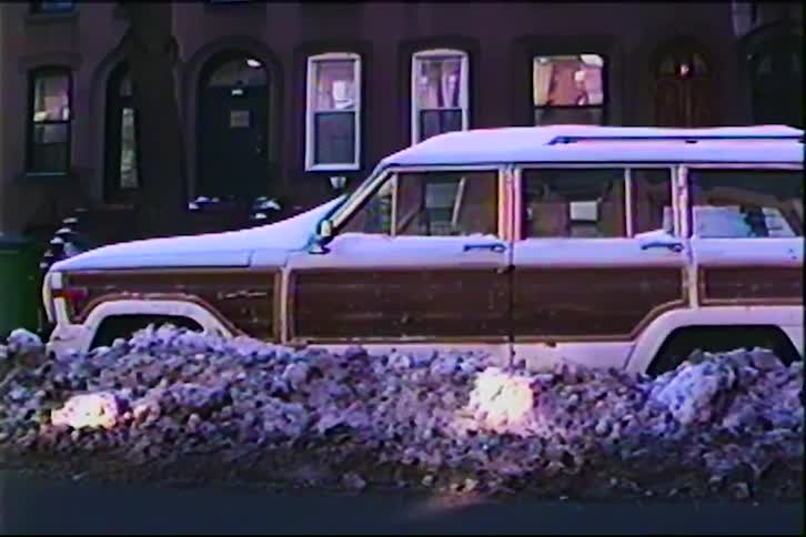 A vehicle is snowed in on a city street in front of an apartment building