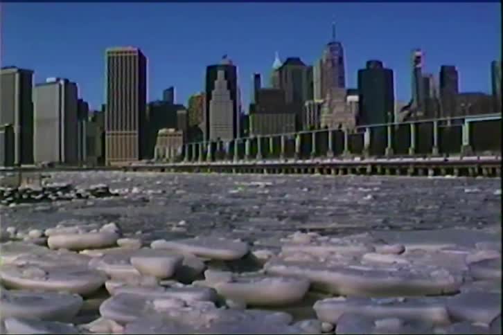 A city skyline rises in the background beyond a waterfront covered with floating chunks of ice under a clear blue sky