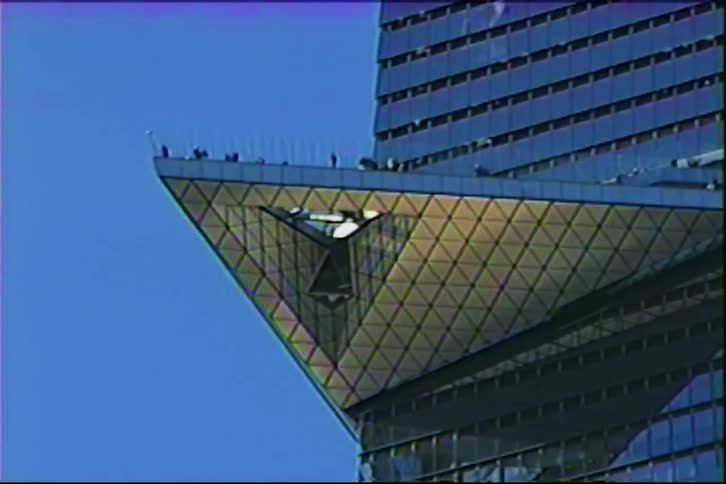 Filmed in the style of a vintage home movie tiny figures of people walk along the edges of a dramatic triangular observation deck jutting out from a glassfaced skyscraper against a clear blue sky