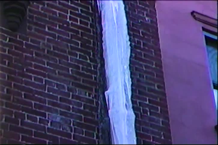 A large white icicle hangs from the side of a red brick building while sunlight creates a warm glow on a nearby wall in a city during winter