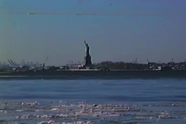 The statue of liberty stands in the distance across a wide expanse of water dotted with floating ice under a clear sky