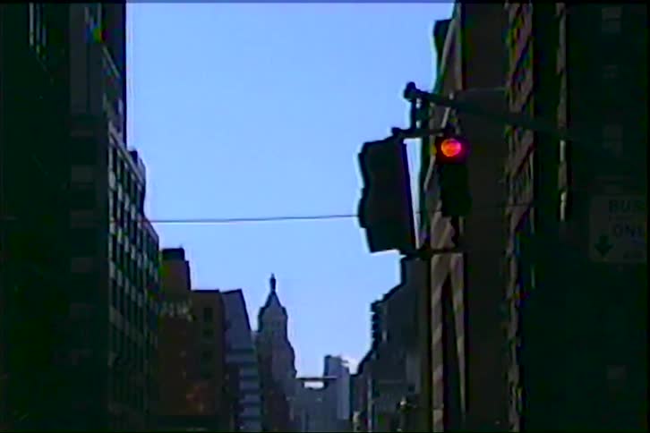 Videoa view looking down a city street shows tall buildings in silhouette while a traffic signal turns from red to green under a clear blue sky