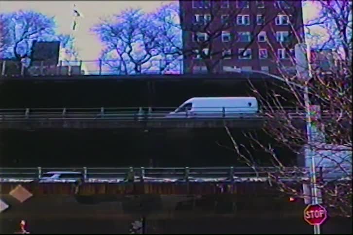 In the style of a vintage home movie several white vans and cars drive across a multilevel urban overpass while a tall brick apartment building and bare winter trees stand in the background