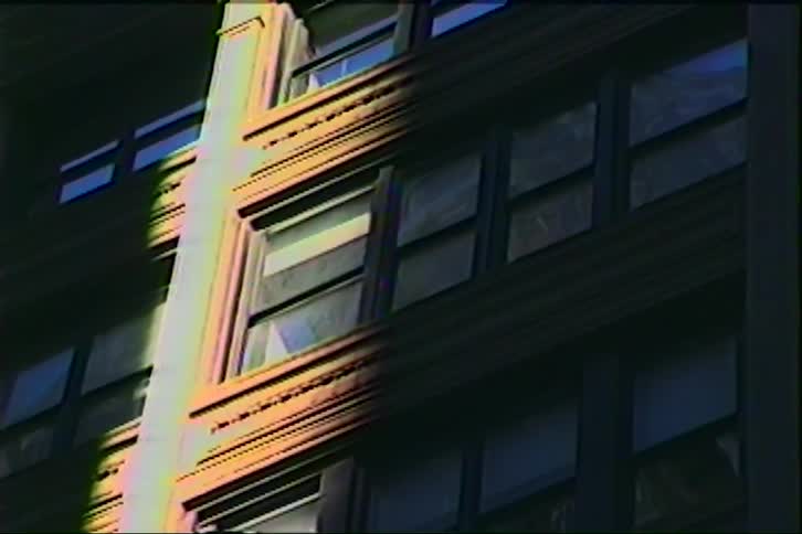 A side view of a dark brick apartment building with many rectangular glass windows features a stripe of warm yellow sunlight and rainbow light leaks across the stone facade in an urban setting