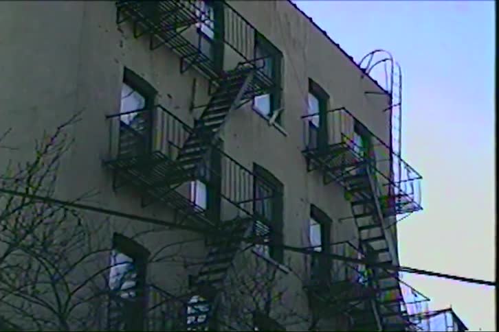 A grainy handheld shot captures the side of an urban apartment building with prominent black metal fire escapes framed by bare tree branches against a pale sky in the style of a vintage home movie