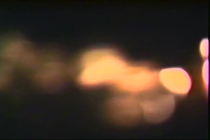 A close view of several small round candles flickering with warm orange light in a dark room shows a grainy film texture and soft bokeh effects while the camera moves slowly toward the flames