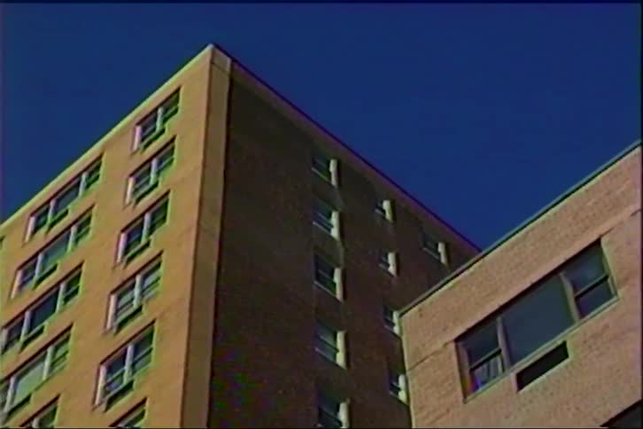 An upward view of several tall brick apartment buildings with many windows under a clear blue sky features a grainy film texture and static distortion typical of an old video recording