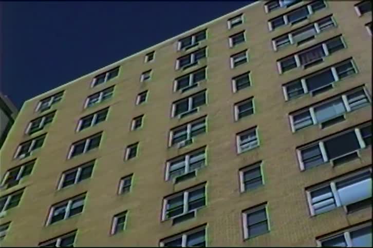 A view of a tan brick apartment building with many rectangular windows and air conditioning units under a clear blue sky features a grainy film texture typical of an old video recording