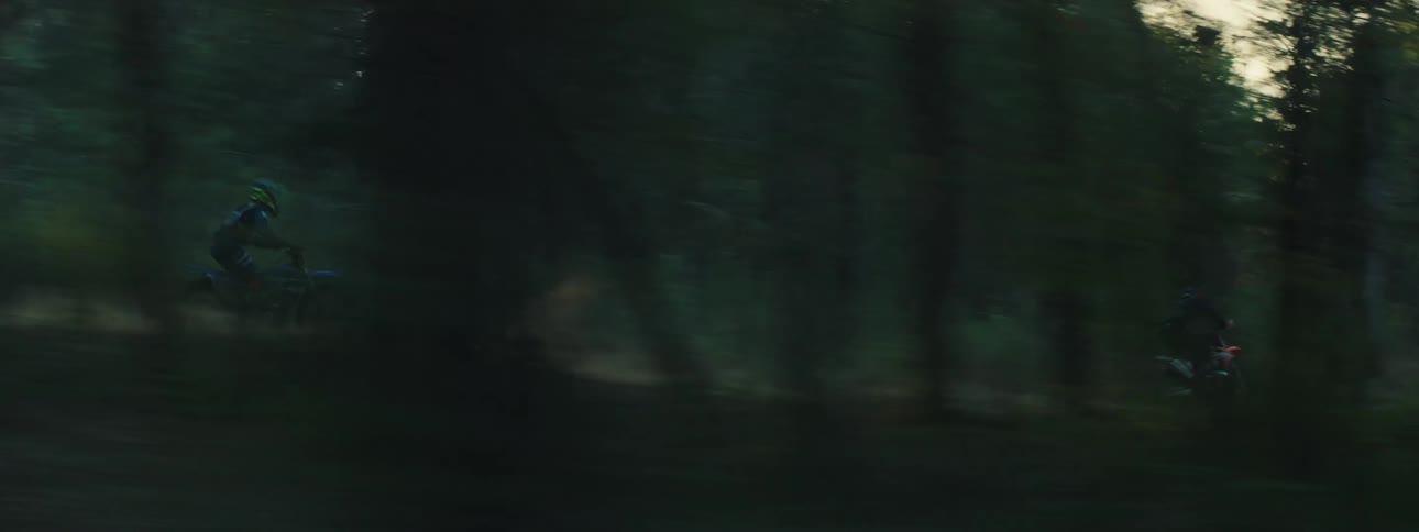 This video features a highspeed tracking shot of several dirt bike riders racing through a dense wooded trail the camera pans rapidly to the left capturing the riders as they kick up dust and navigate between the trees during the late afternoon