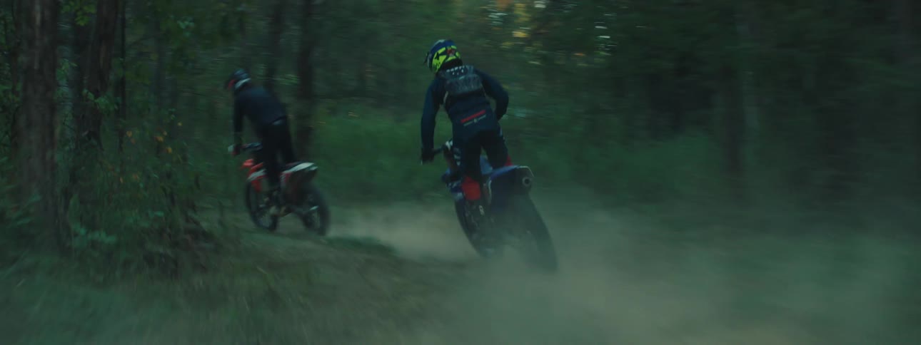 Two motocross riders race along a narrow dirt path kicking up thick clouds of dust as they speed through a dense forestthe camera follows closely behind capturing the highspeed movement and the blurred greenery of the surrounding woods