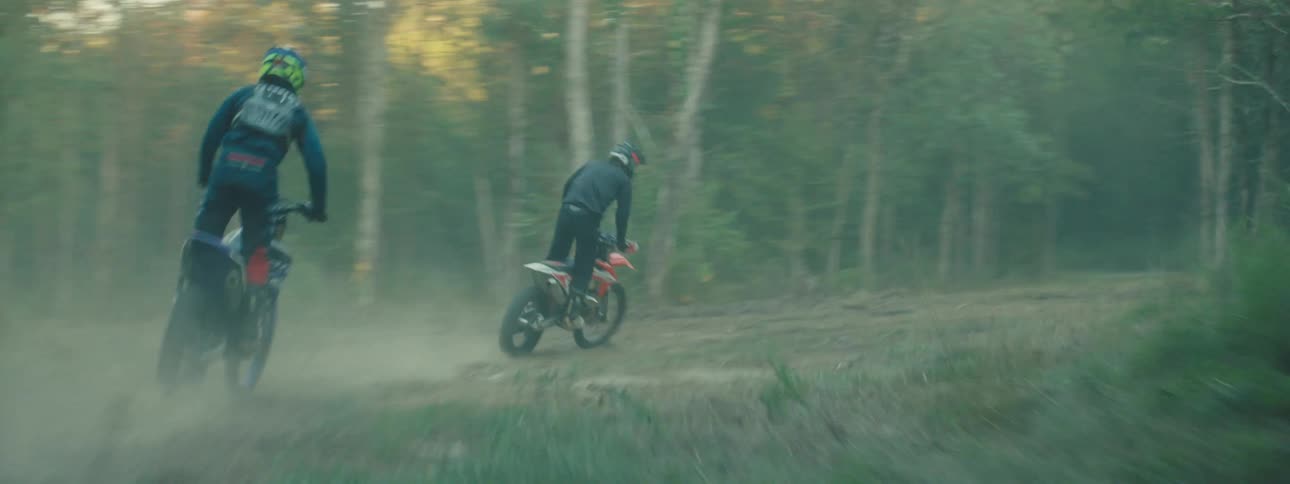 Two people drive dirt bikes along a trail that bends through a forest
