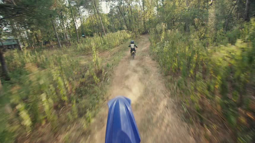 A person wearing a helmet and black gear rides a small yellow motorcycle down a dirt path through a sunlit forest while leaving a trail of dust in a following view