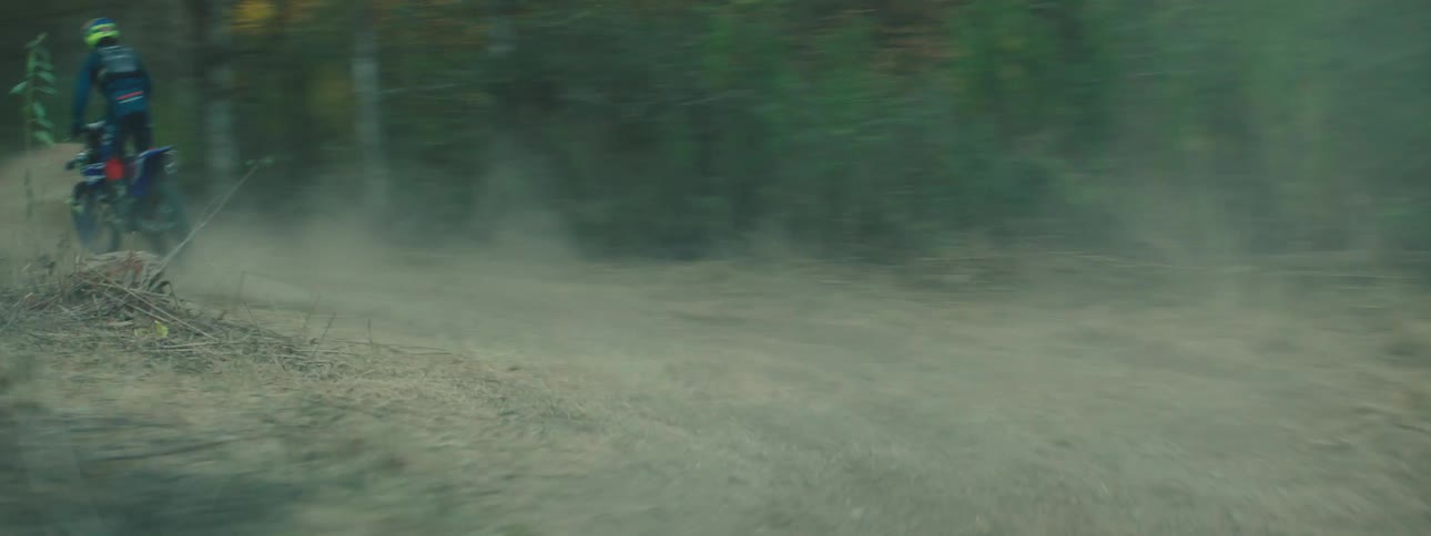 Two dirt bike riders speed along a dusty trail through a wooded area kicking up clouds of dust behind them
