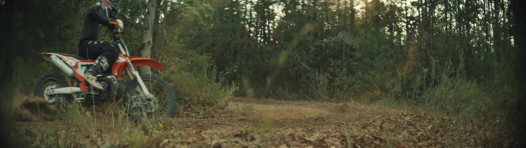 A person wearing a helmet and black gear rides an orange dirt bike through a sharp turn on a wooded trail kicking up a large cloud of brown dust from the ground during a sunny afternoon