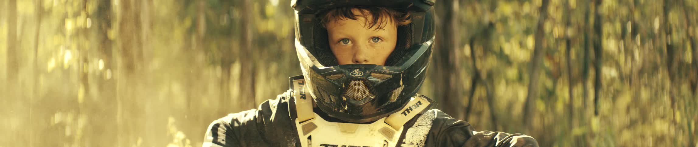 A young boy with bright blue eyes stares intensely into the camera while wearing a black and white motocross helmet and chest protectorthe background is a softfocus forest with sunlight filtering through the trees highlighting the dirt a