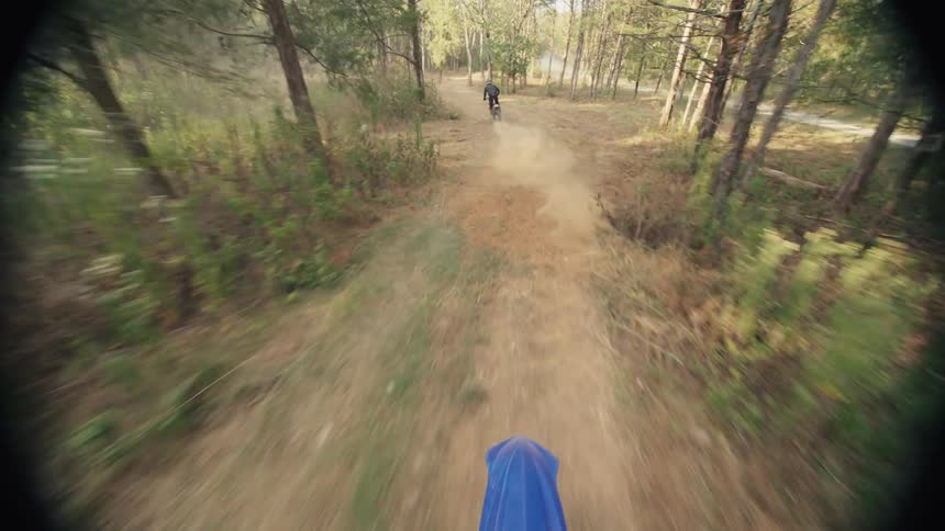 People ride dirt bikes down a trail through the woods across the road from a house on an autumn day