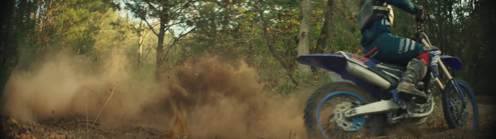 A motocross rider speeds down a sunlit forest trail on a blue dirt bike executing a sharp drifting turn that kicks up a massive swirling cloud of dirt that completely fills the air