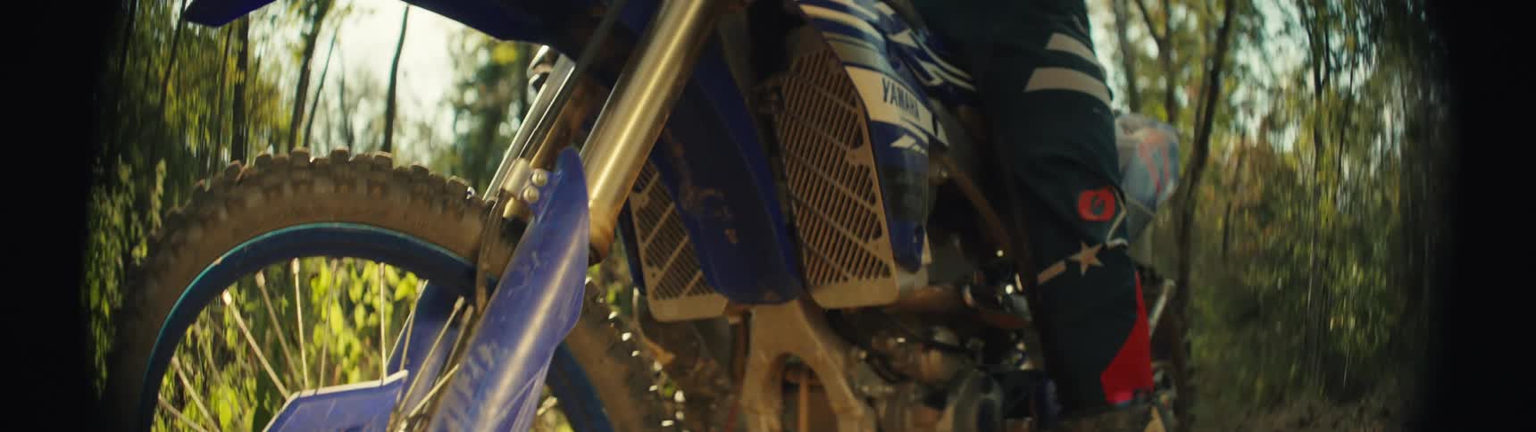 Front wheel of a dirt bike with blue guard rides along a dirt trail under sunlight among trees