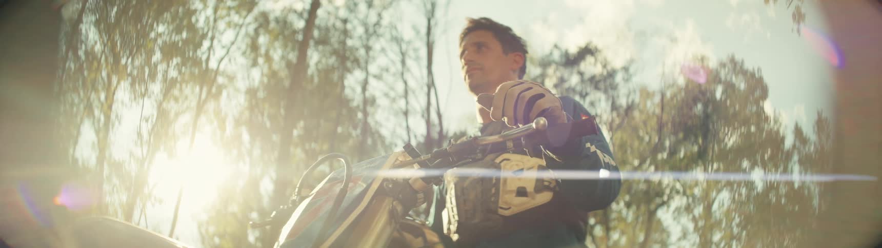 Dirt bike rider holds the handlebars under strong sunlight in a wooded area