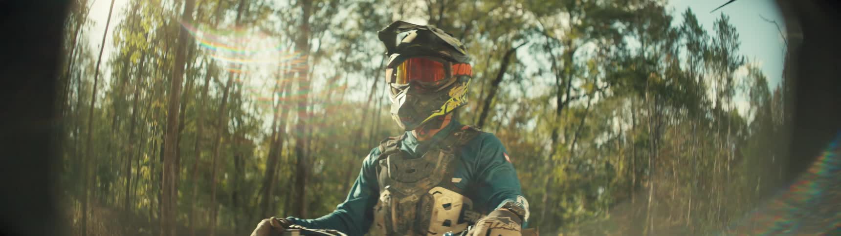 A motocross rider in full protective gear and a helmet sits on a bike in a sundrenched forest the camera slowly pans from a side profile to a frontfacing shot as light flares across the lens