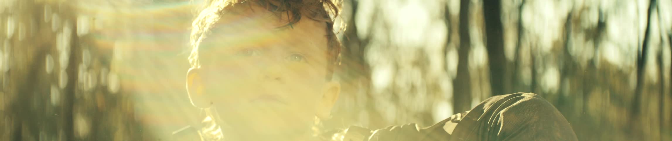The video shows a closeup of a young boy with dirt on his face looking into the distance with a serious expression warm sunlight flares across the lens momentarily obscuring his features before fading as he stares directly toward the camera