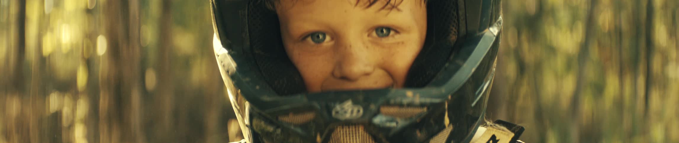 A young rider peers out from beneath a motocross helmet his blue eyes steady in the warm forest light