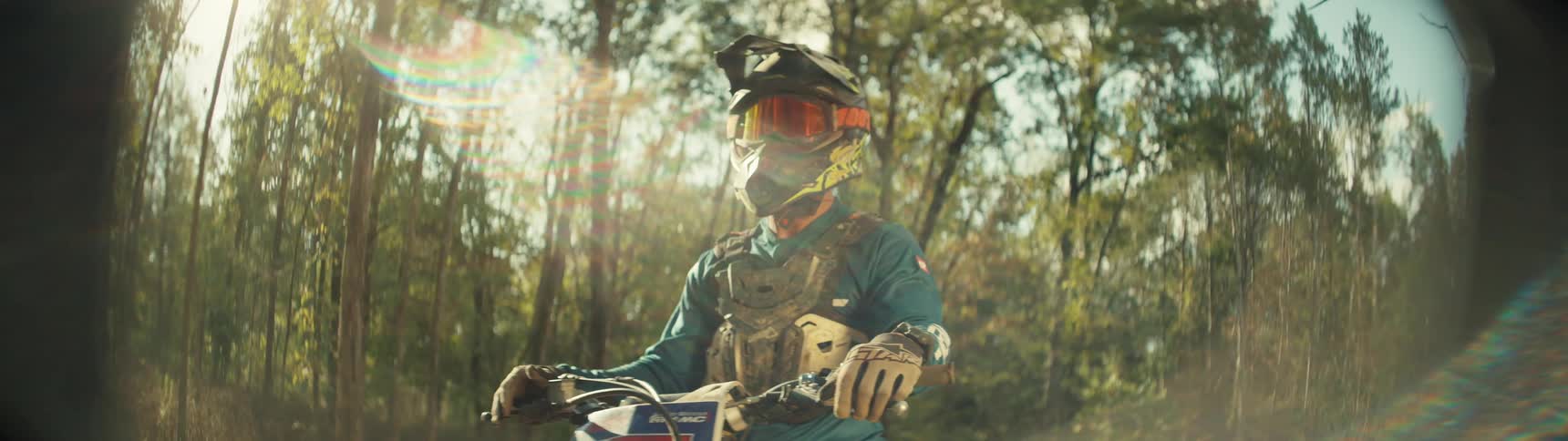 A dirt bike rider sits stationary on their motorcycle fully geared in a helmet goggles and protective chest plate the scene is set in a sundrenched forest where bright lens flares catch the light as the rider looks forward