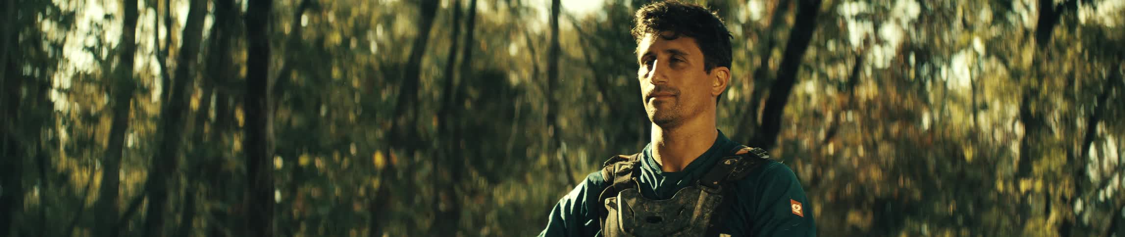 A man wearing a blue shirt and protective body armor stands in a forest with sunlight filtering through the trees in the background