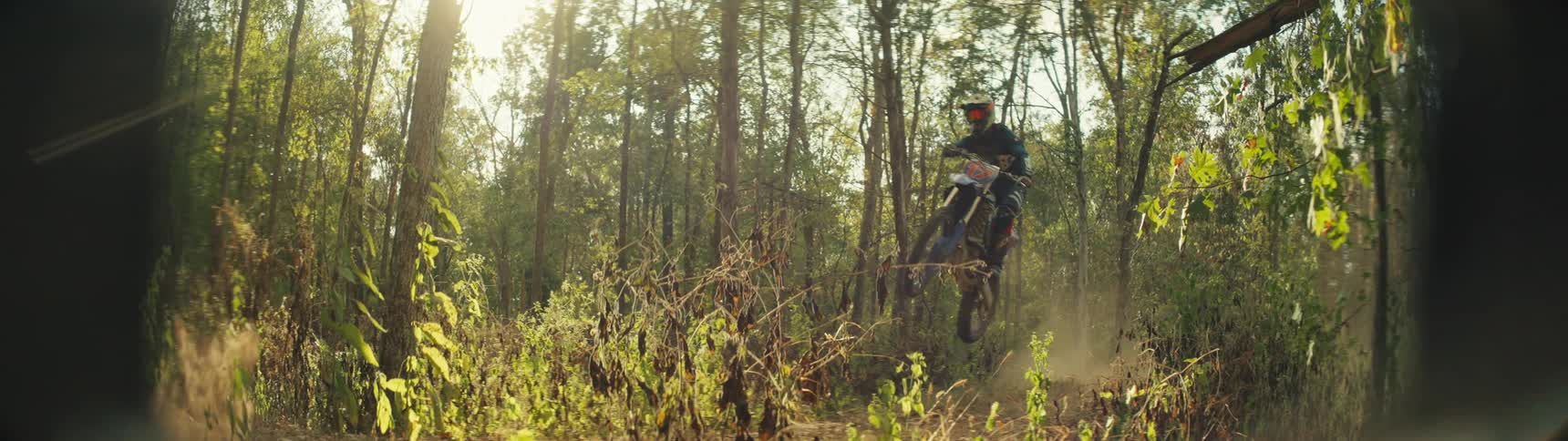 Two men wearing helmets and protective gear jump their motorcycles over a dirt mound in a sunlit forest while leaving a trail of dust in the air
