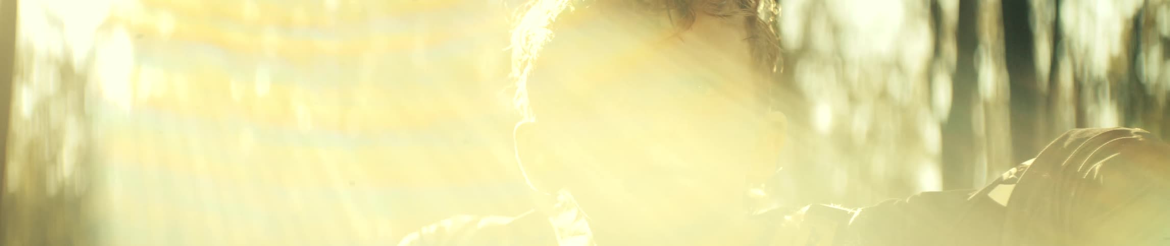 A close view of a young boy with blue eyes and damp hair looking ahead with a serious expression while bright golden sunlight creates a strong glow and lens flares around his face