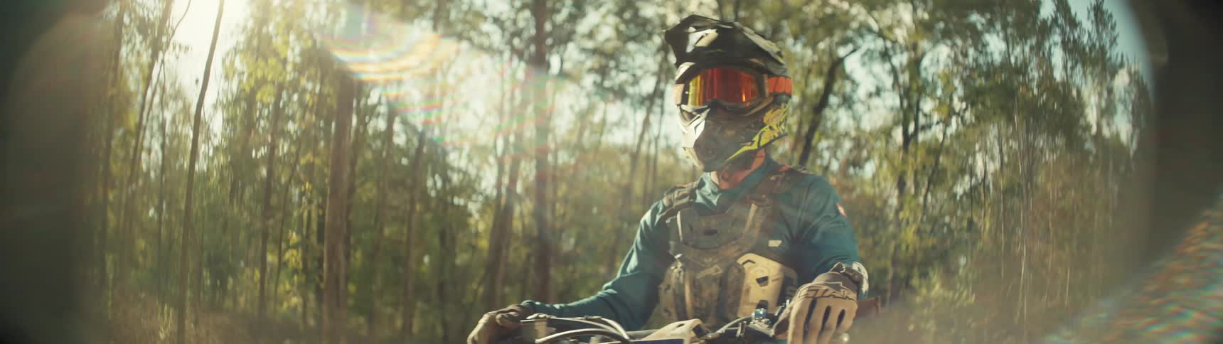 Adult man wearing a helmet and a roost deflector standing with a motorcycle in front of a grove next to a fence outdoors during day time