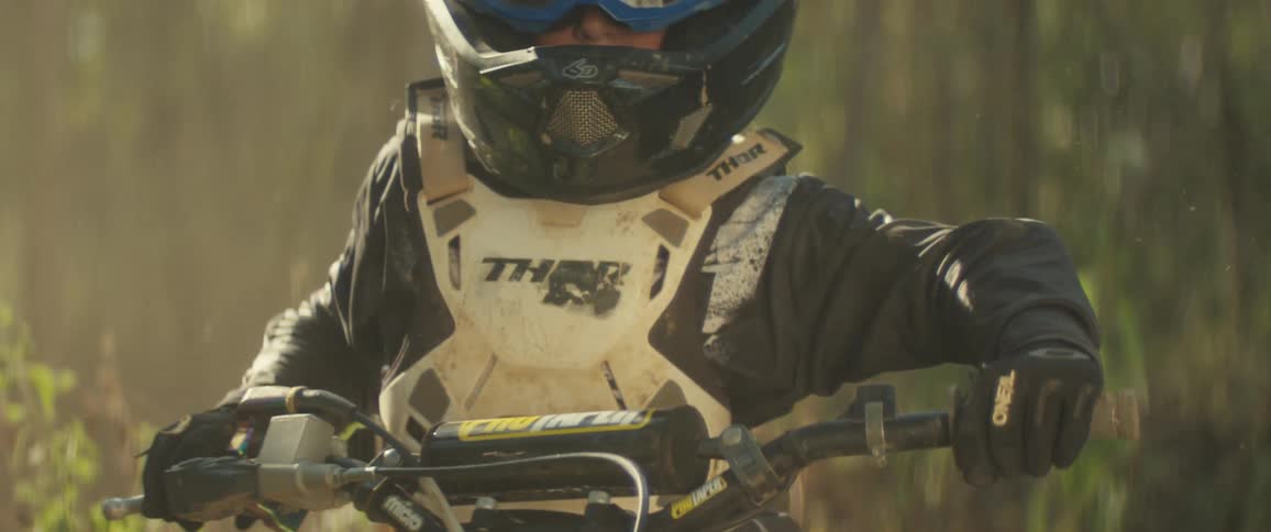 A young boy riding a mountain bike in the woods at midday wearing a black jacket goggles and a helmet