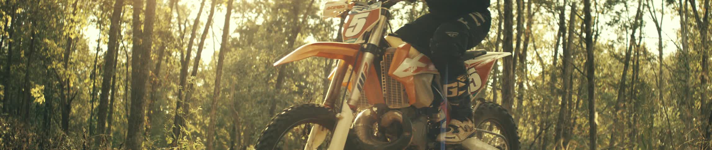 A rider sits astride an orange dirt bike marked with the number 5 sunlight glinting off the frame in the forest