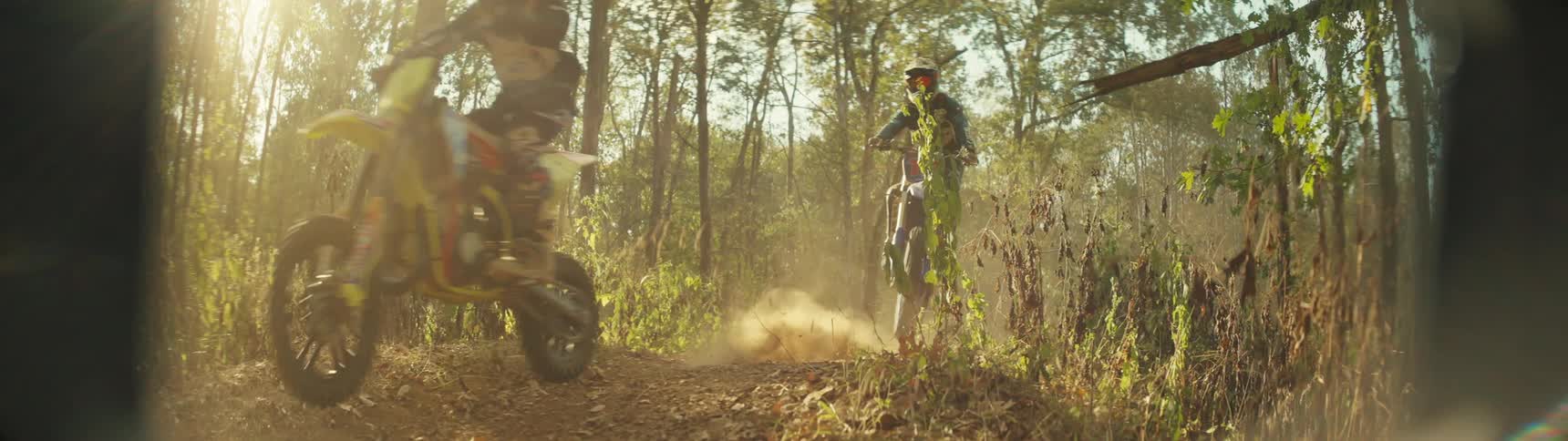 Two dirt bike riders one smaller and one larger speed across a dirt path in a sunlit forest they kick up a thick cloud of golden dust as they ride toward and past the camera