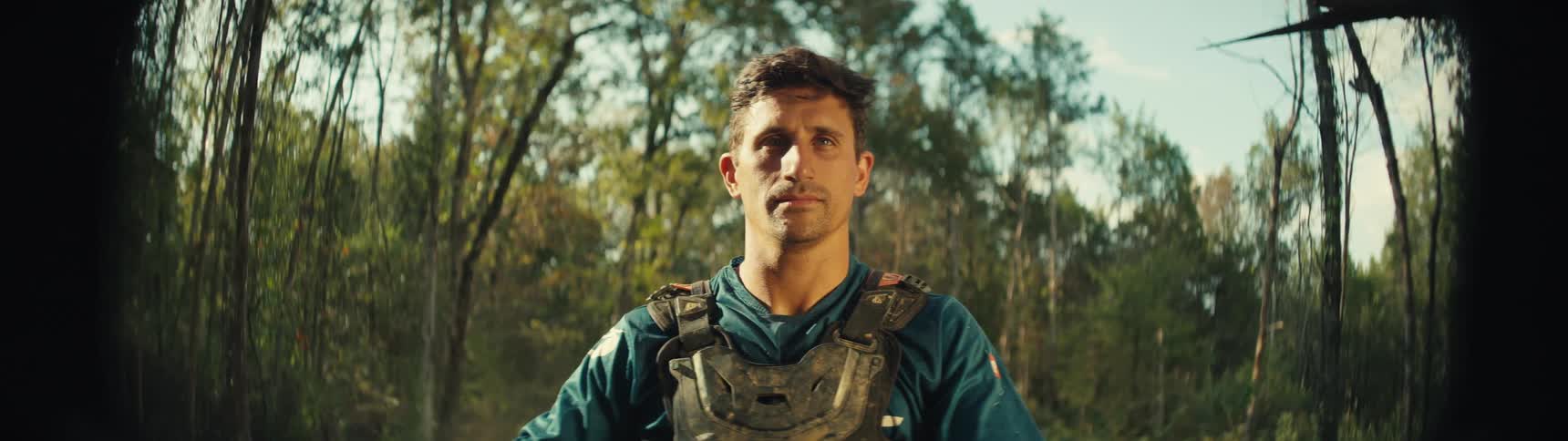 A man wearing dirtsplattered motocross chest protection stands in a wooded area he looks directly into the camera with a calm steady expression while sunlight filters through the trees behind him