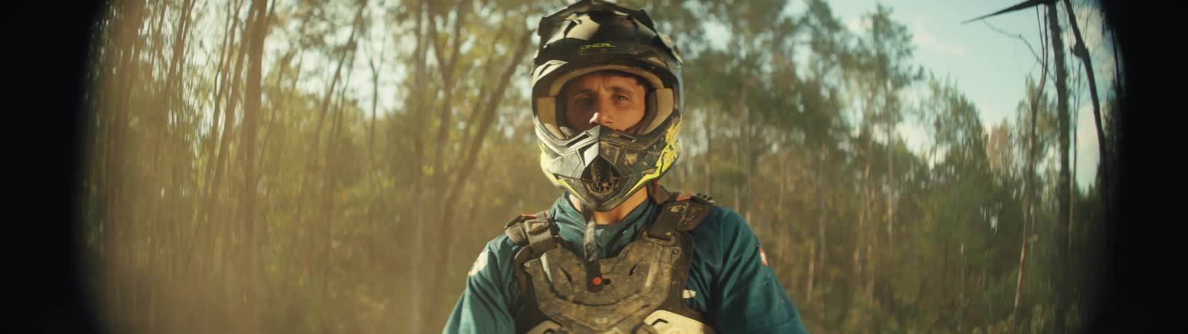A man wearing a black motocross helmet and chest protector stands in a sunlit forest while looking directly at the camera
