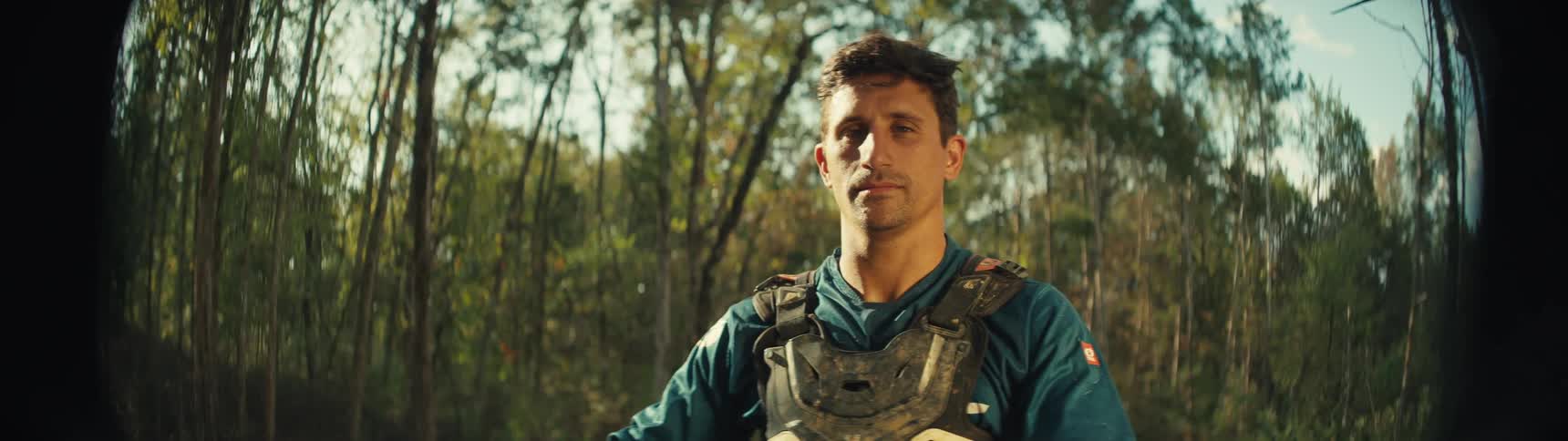 A man in motocross protective gear stands in a sunlit wooded area he maintains a steady serious gaze directly toward the camera throughout the sequence