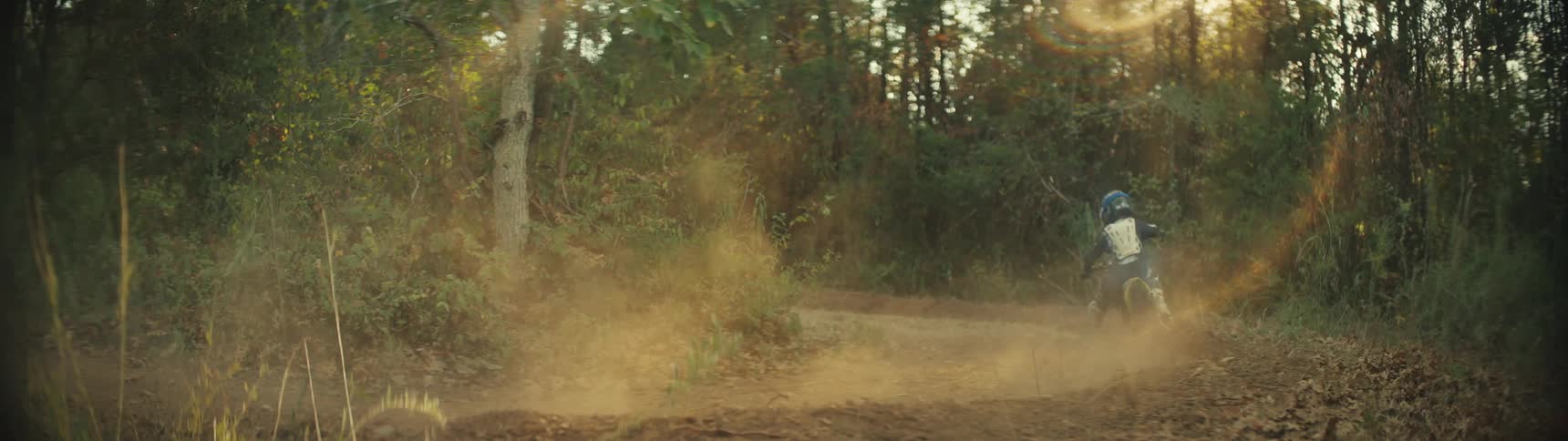 Boy rides a dirt bike around a bend on a forest trail kicking up dust around him