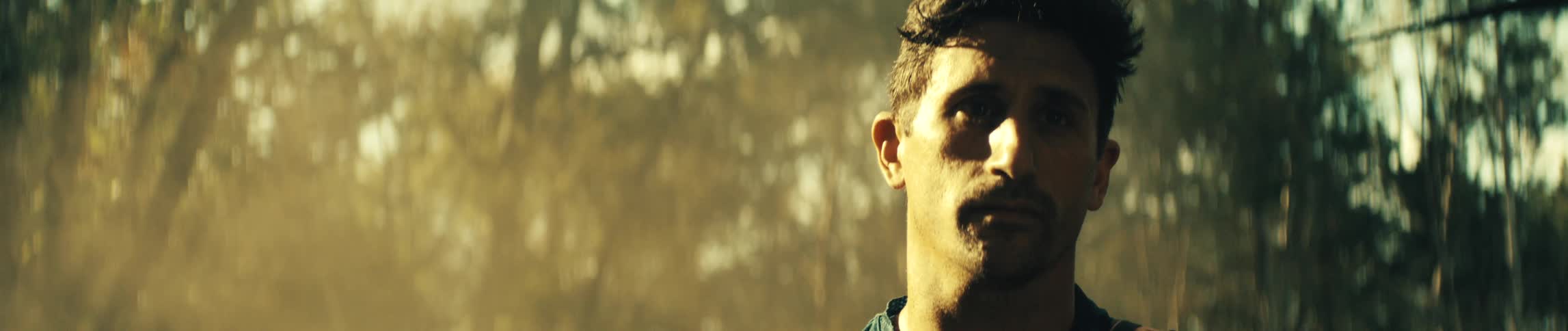 Man with a serious expression looks ahead in an outdoor setting with blurred trees in the background