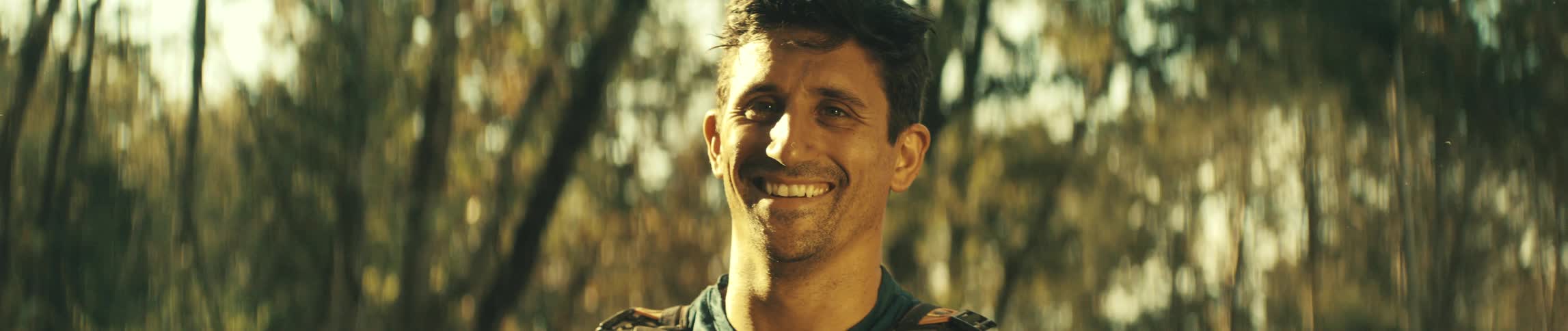 A man with a smile on his face looks directly at the camera in an outdoor setting