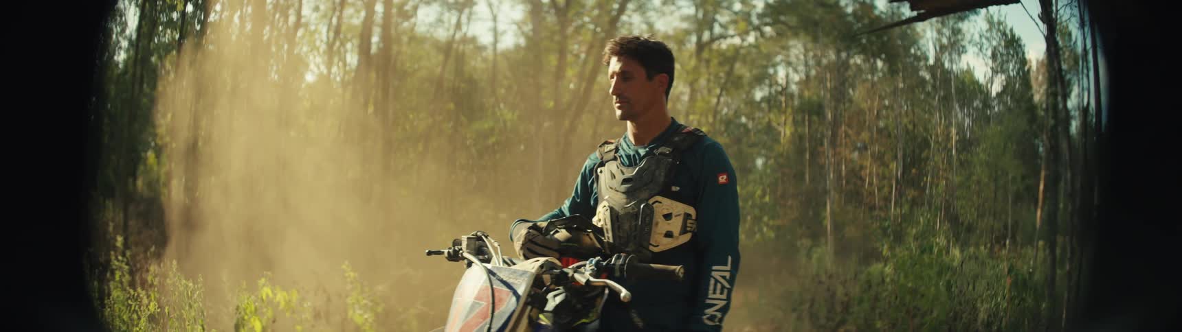 Man stands next to a dirt bike on a forest trail with dust around him