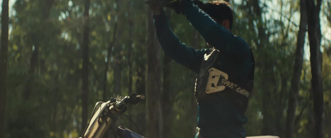 A young male mountain biker in the woods wearing a blue jersey takes off his helmet at sunset